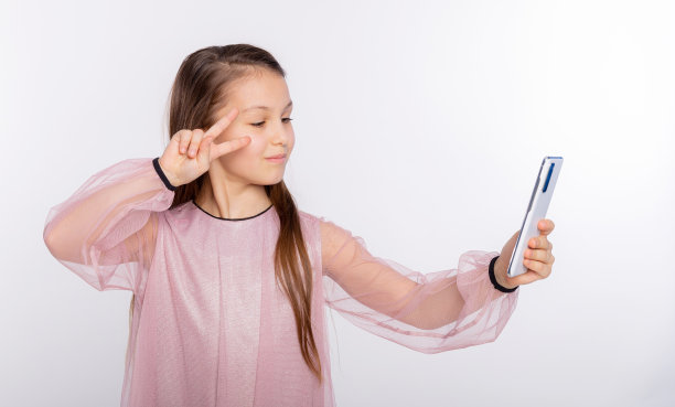 身穿粉色连衣裙的小女孩对着智能手机的摄像头飞吻，并在白色背景上自拍。为你的文本腾出空间图片下载