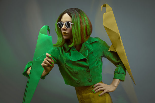 一个美丽的年轻时尚模特的肖像戴太阳镜，一件绿色的夹克和一个绿色的波波头，摆姿势与两只折纸鹦鹉前面的灰色背景图片下载