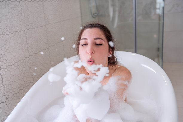 年轻的千禧一代女性，头发盘成一个髻，正在洗澡，玩得很开心，吹着她手上的泡沫图片下载