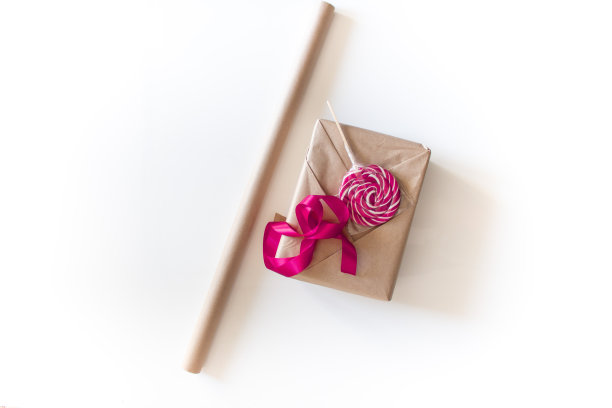 节日礼品包装。盒子用米色牛皮纸包裹着，旁边放着一根粉色的丝带和一个圆形的棒棒糖作为装饰。把情人节礼物包装成白色背景的过程图片下载