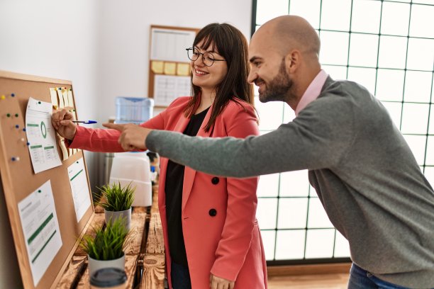 两名西班牙裔企业员工微笑着在办公室的软木板上写着提醒。图片下载