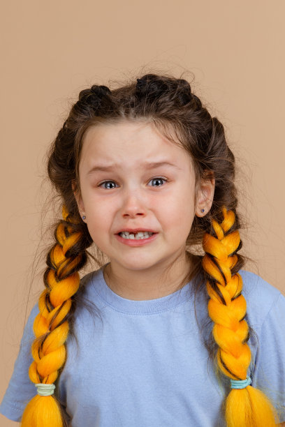 心烦意乱的小女孩张开嘴，眼泪在眼睛哭，头上扎着黄色的kanekalon辫子，穿着浅蓝色的t恤，米色的背景图片下载