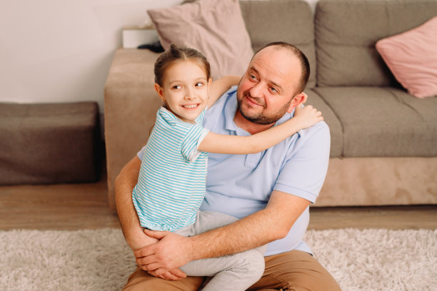 近距离的照片，爸爸和他的可爱的小女孩周末在家花时间坐在地毯上拥抱和看着一边。图片下载