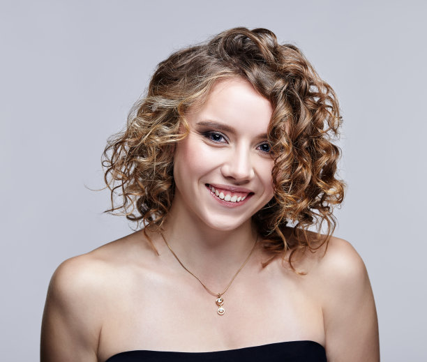 年轻微笑的女人与卷曲的头发在灰色的背景美丽的肖像。图片下载
