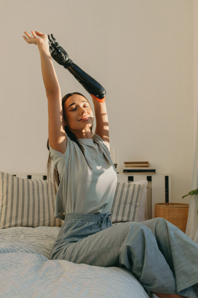 早晨，有吸引力的温柔的美丽的照片年轻的女孩，20岁伸展她的背部，举起手，坐在床上，在窗户前，向太阳致敬，手臂上有仿生假肢图片下载