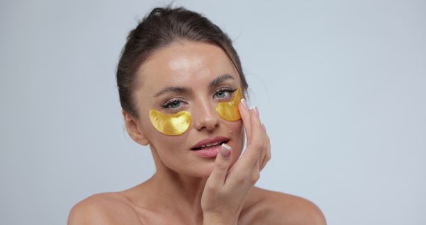 女人的肖像与金眼罩和触摸她的脸，而看着相机。裸肩和化妆的女性摆姿势微笑。美容治疗。图片下载