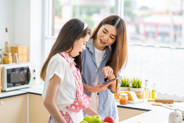 在厨房里，年轻的亚裔妹妹一边用手机寻找食谱一边聊天。移动智能手机技术与人们的生活方式。素材图片