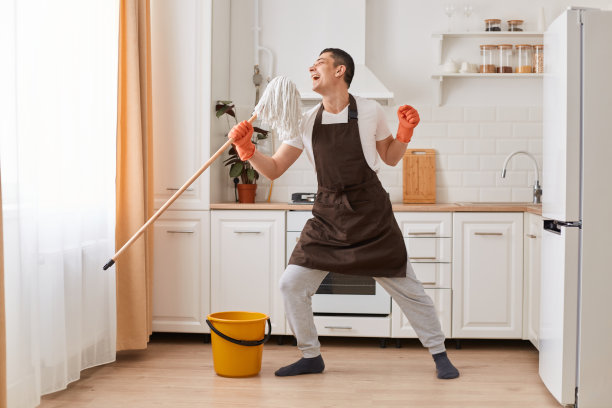 穿着棕色围裙的白人男子兴奋而快乐地在厨房洗地板，用拖把唱歌，表达积极的情绪的全身肖像。图片下载