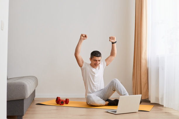 室内拍摄的这张照片显示，一名身穿白色t恤和灰色裤子的男子非常兴奋，坐在笔记本电脑旁的地板上，举起双臂，握紧拳头，庆祝锻炼结束。图片下载