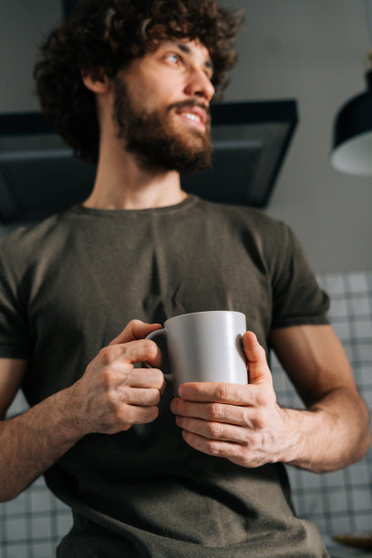 近距离低角度的视角，沉思的年轻人手中握着一杯早上的咖啡，沉思地看着别处，站在厨房里图片下载