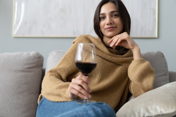 漂亮、迷人、时尚的年轻女子坐在沙发上举起了红酒酒杯，审视着，品尝着饮料图片下载