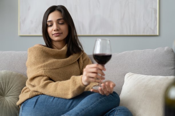 漂亮、迷人、时尚的年轻女子坐在沙发上举起了红酒酒杯，审视着，品尝着饮料图片下载