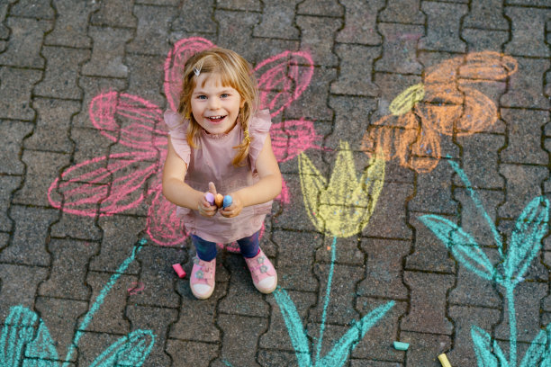 学前班的小女孩用彩色粉笔在后院的地上画花。积极快乐的蹒跚学步的孩子在沥青上画画和创作图片。夏季儿童创造性户外活动。图片下载