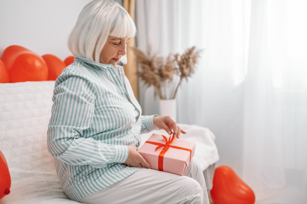 一个40到50岁的金发女人，穿着时髦的衬衫，坐在家里的沙发上打开一个带有红色丝带蝴蝶结的惊喜礼盒。庆祝情人节。母亲的一天图片下载