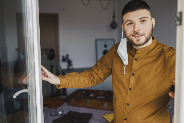 一个戴着口罩的年轻人坐在隔离室内，从窗户往外看图片下载