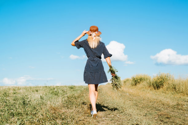 在阳光明媚的日子里，优雅美丽的年轻女子穿着夏装，戴着草帽漫步在大自然中，背景。白种人金发女孩带着一束野花走在户外的田野图片下载