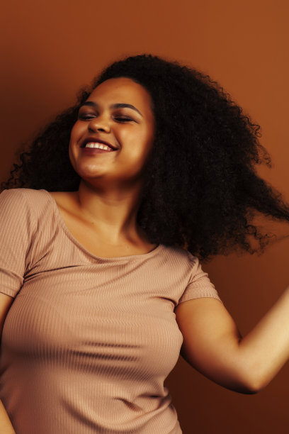 漂亮的年轻非洲裔美国妇女与卷曲的头发摆愉快的手势在棕色的背景，生活方式的人的概念图片下载