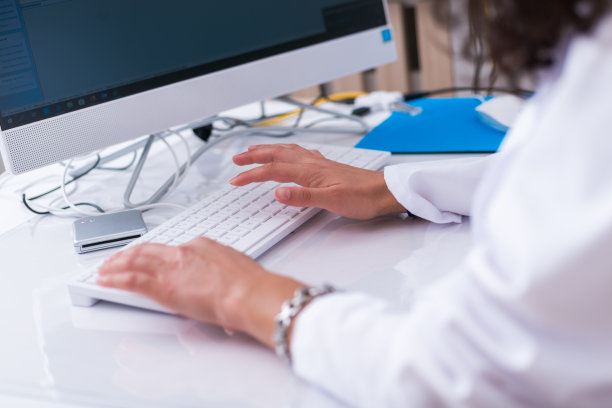 靠近一位女医生(护士)坐在医院大厅的白色桌子上敲击白色键盘的双手。图片下载