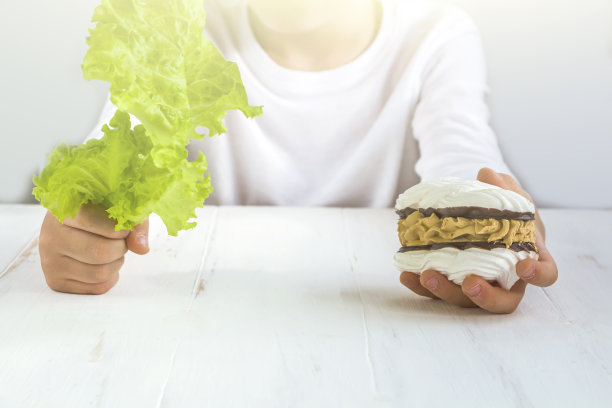 不健康食品vs健康食品。小男孩手里拿着蛋糕和生菜。孩子在糖果和蔬菜之间做选择。模糊，选择性的聚焦食物，背光图片下载