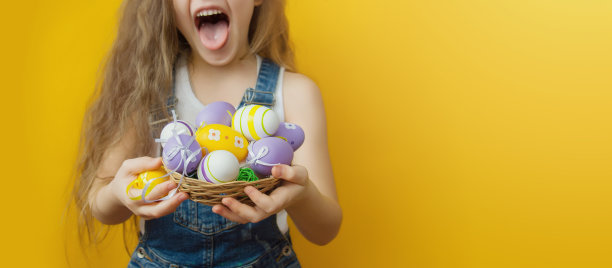一个调皮的小女孩穿着白色t恤和牛仔工作服，黄色的背景上，她手里拿着一个小小的柳条篮子，篮子上放着复活节彩蛋。有选择性的重点。特写镜头。Copyspace。布局广告。素材图片