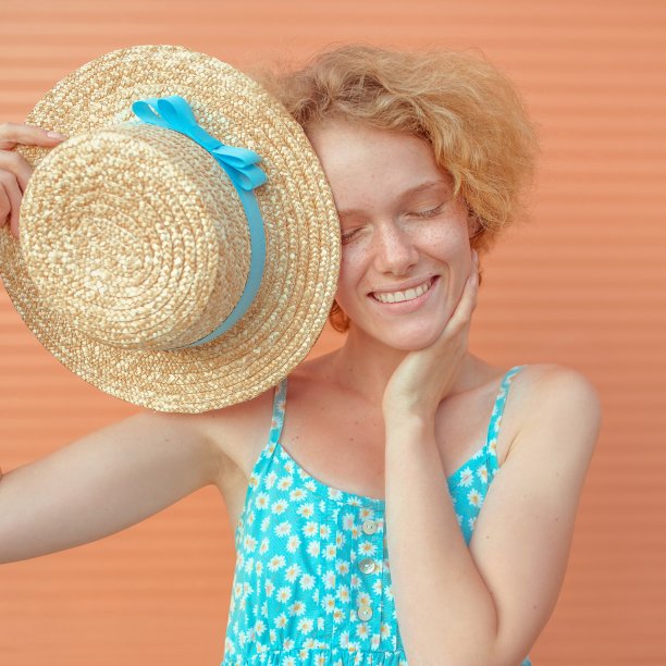 年轻活泼的鬈发女子，身穿蓝色背心裙，手里拿着米色背景的草帽。趣味、夏日、时尚、青春的理念图片下载
