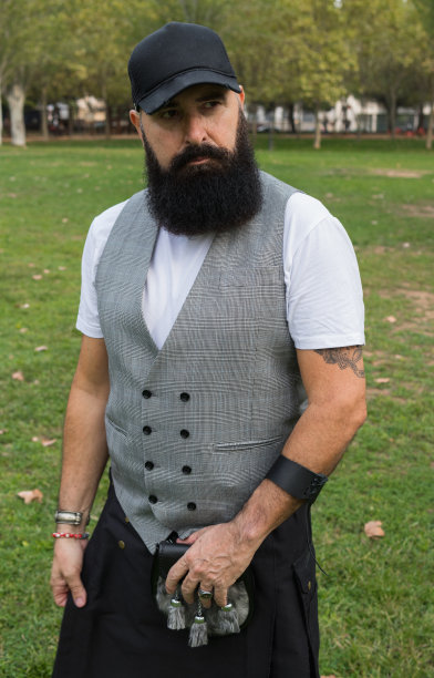 一个留着胡子的时髦男人，穿着短裙、帽子和马甲，站在草地上图片下载