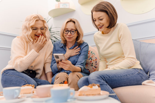 三个有吸引力的年轻女士的肖像看着与喜悦的电话。坐在舒适的沙发上欣赏。图片下载