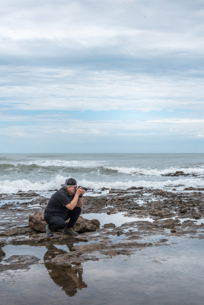 戴眼镜的成年男子跪在岩石上拍照。背景是大海。图片下载