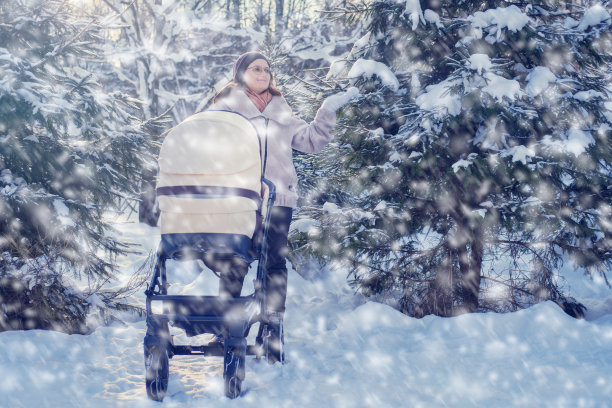 冬天的公园里，白雪覆盖着圣诞树，一位母亲推着婴儿车带着孩子。妈妈推着婴儿车在森林里白雪覆盖的树林里图片下载