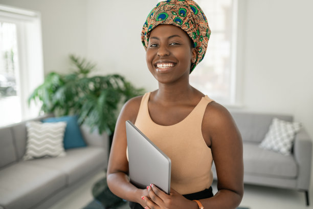 一名非洲妇女坐在家里，微笑着拿着笔记本电脑，戴着头巾图片下载