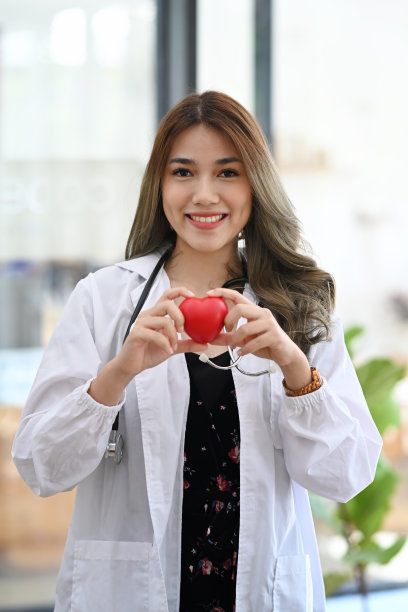 身穿白色制服的女医生拿着红心，对着镜头微笑。医疗保健和心脏病学概念。图片下载