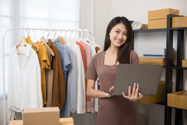 年轻美丽的亚洲女性商人在时尚商店持有笔记本电脑检查库存在轨道服装。在服装店工作的亚洲女性企业家。图片下载