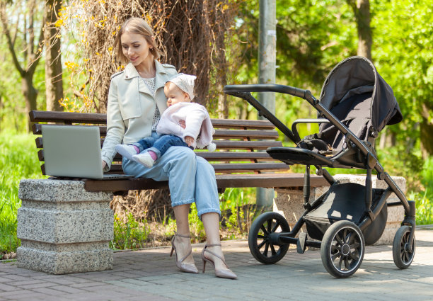 时尚的商务妈妈抱着婴儿坐在公园的长椅上使用笔记本电脑。远程工作，母亲，产假工作图片下载