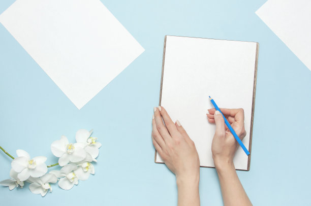 女人的手用温柔的指甲握着铅笔，空白的白纸纸，蝴蝶兰花枝在蓝色的背景上俯视图平躺着。计划的概念。为您的设计提供模型图片下载