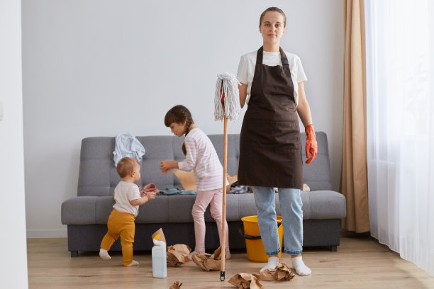 穿着白色t恤，棕色围裙和牛仔裤，面带微笑的积极女性，正在打扫房子，和她的孩子们摆姿势，手里拿着拖把洗地板，看着镜头。图片下载