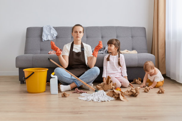 穿着棕色围裙、牛仔裤和白色t恤的妇女正在打扫她的房子，孩子们正在弄得一团糟，她疲惫不堪，试图通过瑜伽练习来放松自己。图片下载