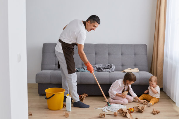 室内拍摄的深色头发的男子穿着休闲的衣服和围裙，洗地板，与他的孩子们摆姿势，手里拿着拖把，看着孩子们和房间里的混乱。图片下载