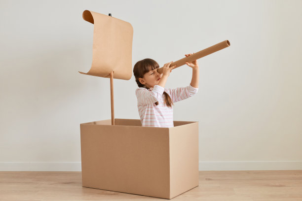 黑头发的小女孩用纸箱做船的画像，拿着望远镜，聚精会神地望着远方的地平线，在家里玩水手游戏。图片下载