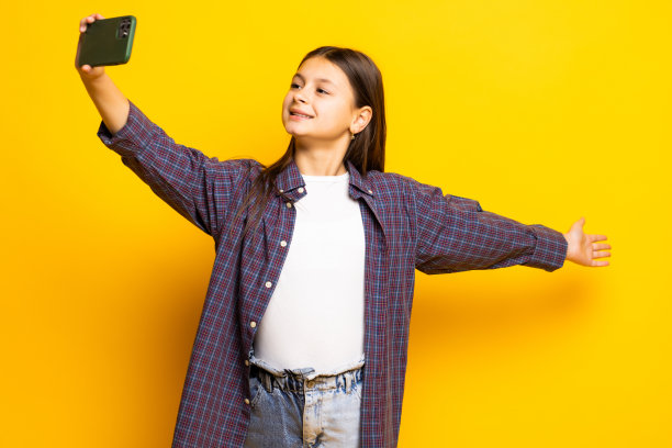 黄色背景下的小女孩手持现代手机，在视频通话中挥动手掌表示你好。人的现代科技观念图片下载