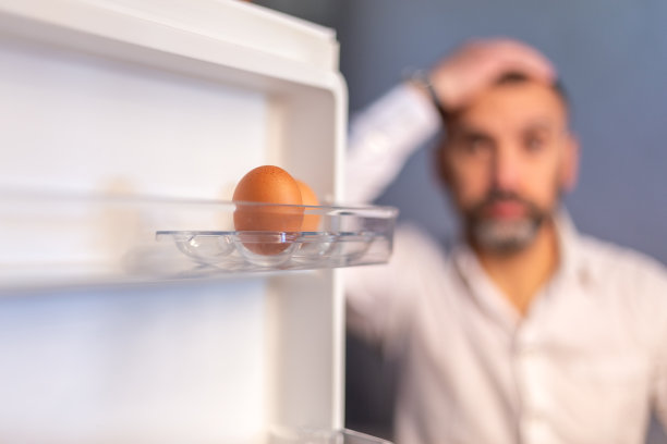冰箱门上有两个鸡蛋。背景中，一名心不在焉的男子盯着空无一物的冰箱。概念:空冰箱。没有吃的吗?图片下载