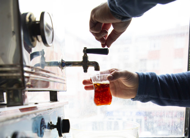 把茶从一个大茶壶倒进茶杯里。公共空间用钢制柴火锅炉。选择性地聚焦红色玻璃图片下载