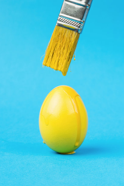 黄色的颜料从黄色的画笔滴到蓝色背景上的黄色鸡蛋上。复活节会议的概念。图片下载