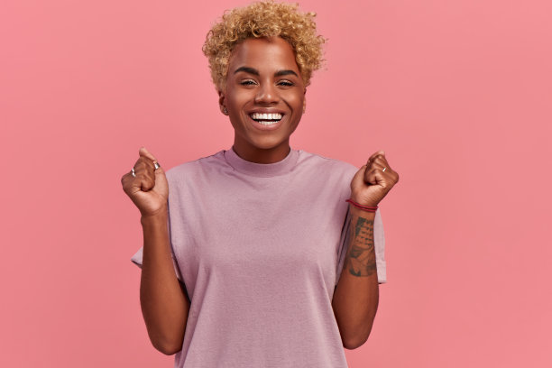 照片中，一名黑人女性开心地握紧拳头，大张着嘴大声呼喊，庆祝自己的成功，在粉红色的背景下摆出各种姿势。快乐的非洲女人，快乐的表情图片下载