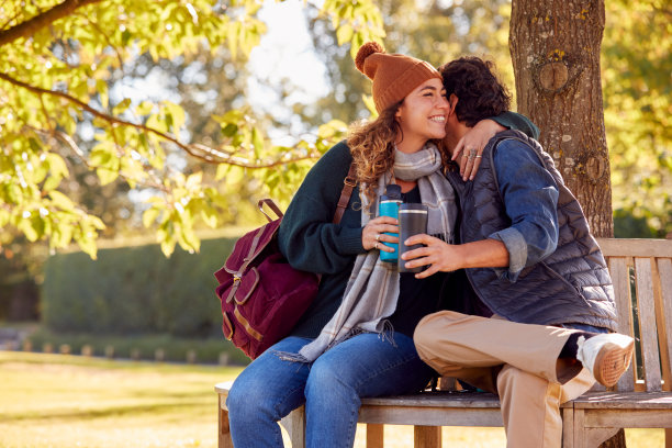 在秋天公园的长椅上，一对夫妇抱在一起，手里拿着可重复使用的杯子图片下载