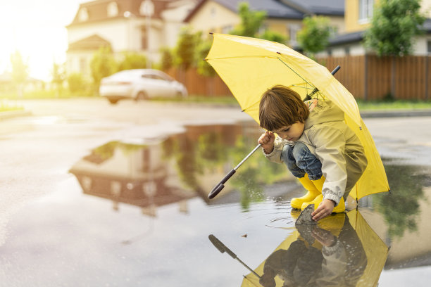 一个孩子撑着一把黄色的伞，站在外面的水坑里。在阳光明媚的日子里，快乐的孩子在房子附近散步，玩耍，玩得开心图片下载