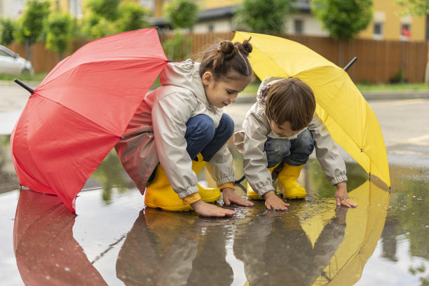 雨后，快乐的孩子们在水坑里玩耍。哥哥和姐姐躲在伞下摸水。孩子们在街上玩得很开心图片下载
