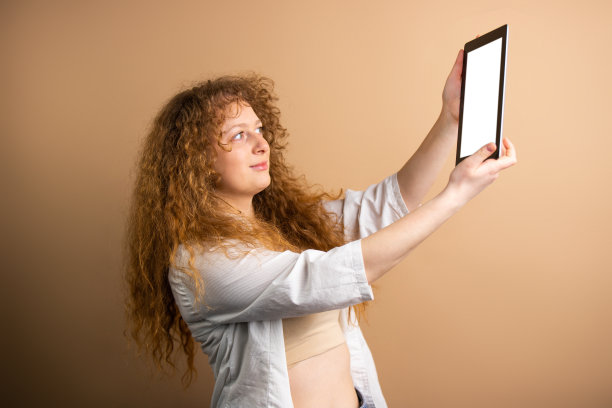 侧视图照片，一个年轻的红发女人手里拿着平板电脑，准备在金色的背景上自拍。图片下载