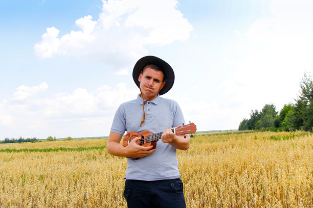 艺术家。年轻漂亮的白人男子在外面弹尤克里里。帅气的嬉皮士在自然背景下弹尤克里里吉他。戴着牛仔帽的男人微笑着弹吉他图片下载