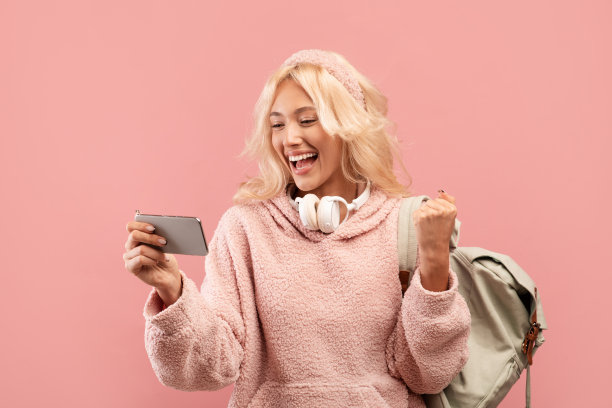 一名女学生手持智能手机，高兴地挥舞着拳头，背着双肩包在粉红色的背景下摆出姿势图片下载