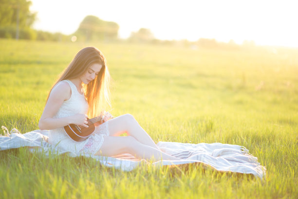 一个女孩在阳光明媚的日子里坐在格子布上，弹着尤克里里琴，在绿油油的草地上谱曲，并在笔记本上写下歌词图片下载